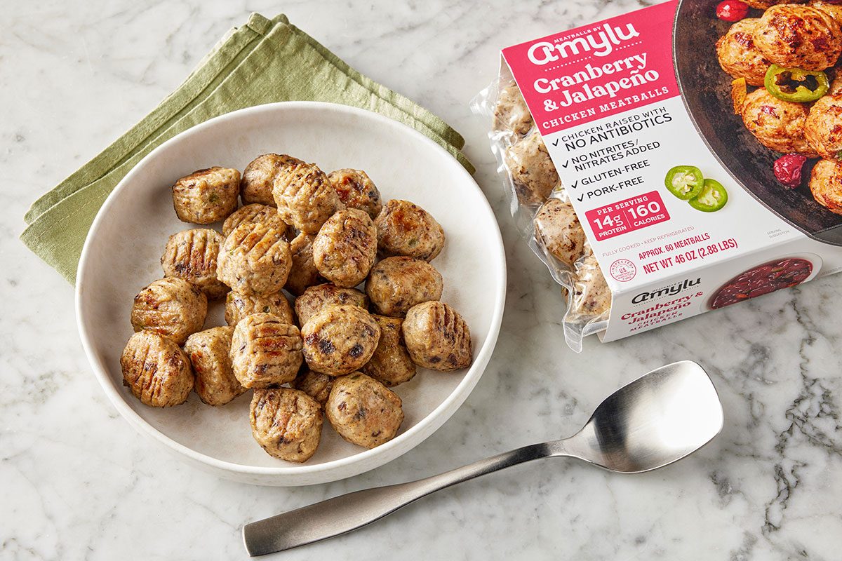 Overhead shot of a white bowl filled with grilled chicken meatballs on a marble surface; Beside it are a package of Amylu Cranberry & Jalape&ntilde;o Chicken Meatballs, a folded green napkin, and a silver spoon