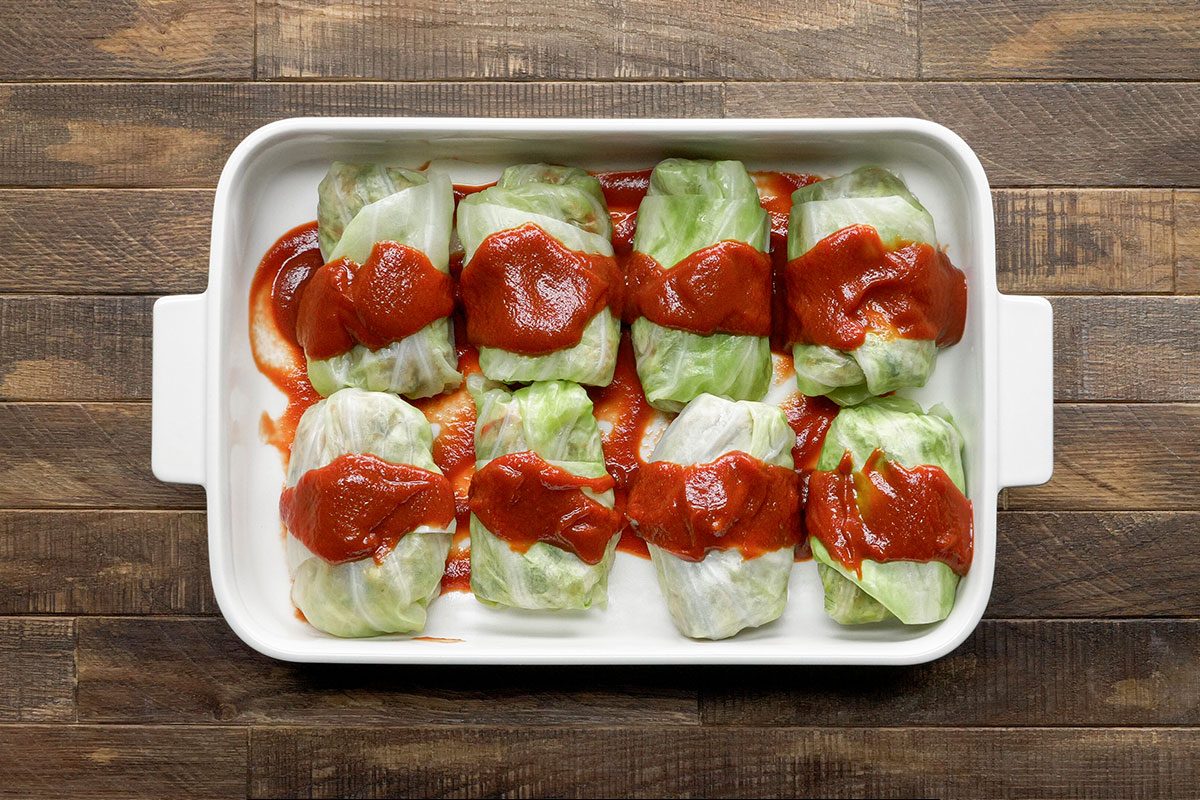 A white baking dish holds eight cabbage rolls topped with tomato sauce, arranged in two rows on a wooden surface.