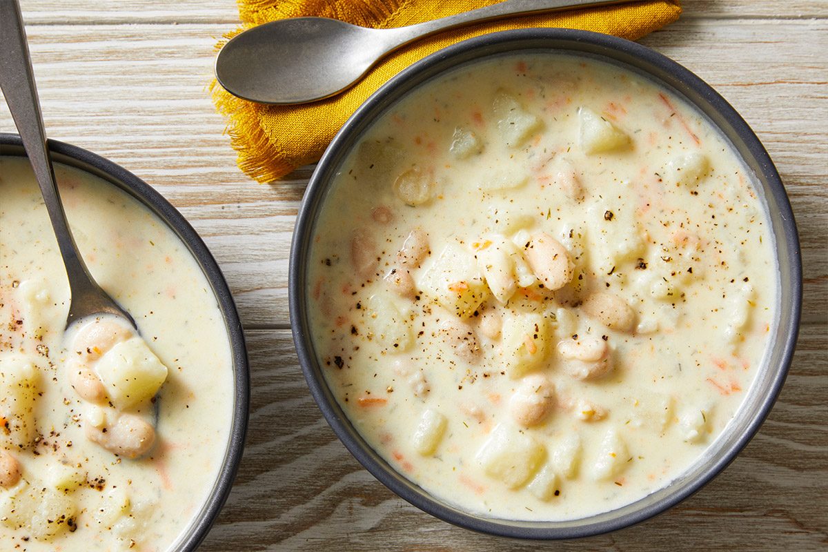Overhead shot of two bowls of Potato Soup with Beans, featuring chunky potatoes and beans topped with black pepper, with spoons and a yellow napkin nearby;