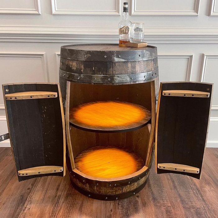 A wooden barrel converted into a storage cabinet with two shelves and open doors. Two bottles and two glasses sit on top. The cabinet sits on a wooden floor against a paneled wall.