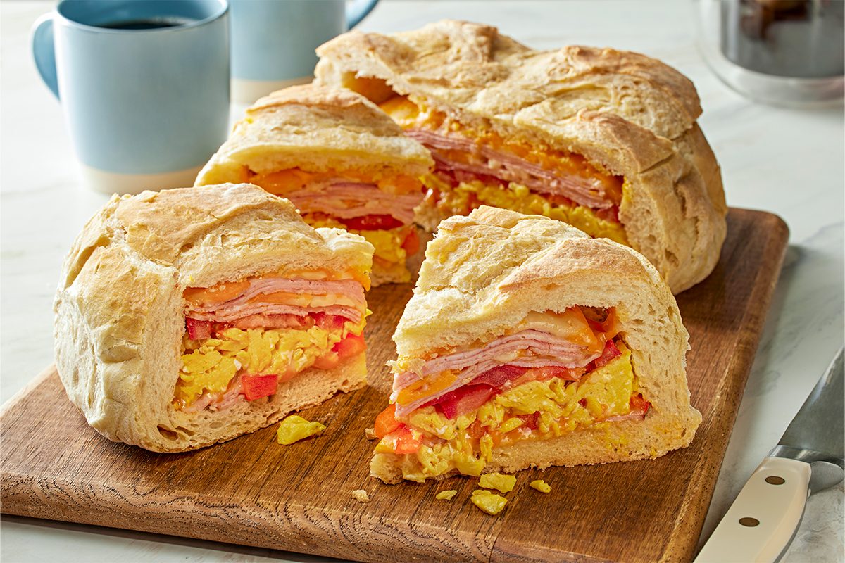 Close shot of a sliced breakfast loaf revealing layers of scrambled eggs, diced peppers, melted cheese, and thinly sliced ham inside a golden, crusty bread round; The pieces sit on a wooden board with coffee mugs and a knife in the background;
