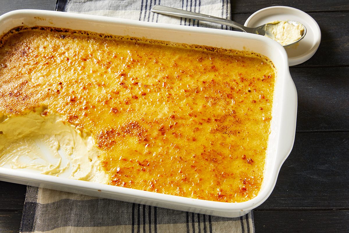 A rectangular casserole dish filled with a golden-brown baked custard dessert, with a portion scooped out, sits on a striped cloth next to a small dish of vanilla powder and a spoon.