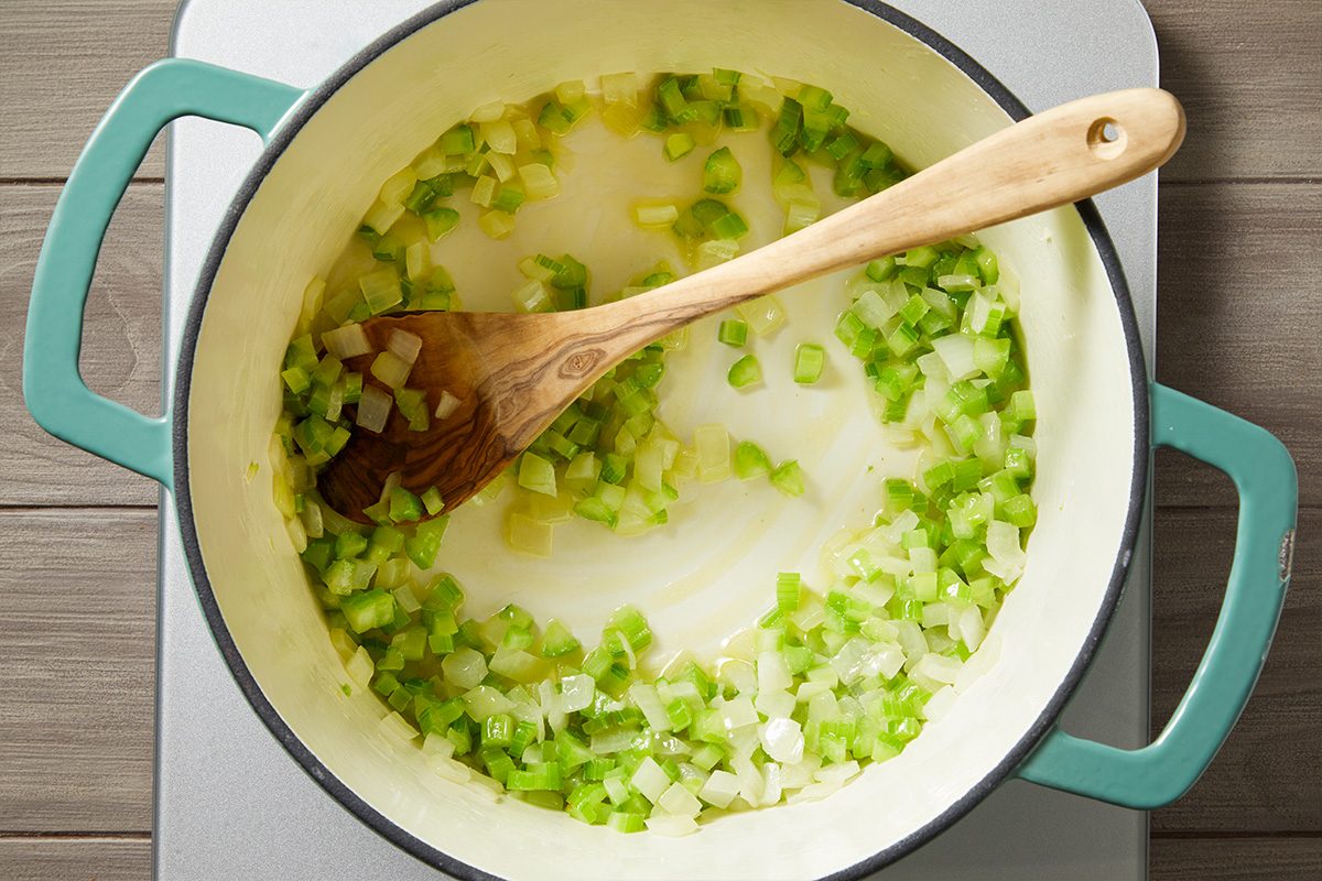 Overhead shot of chopped onions and celery being sautéed in a large light-colored pot with a wooden spoon, as the pot sits on a stovetop