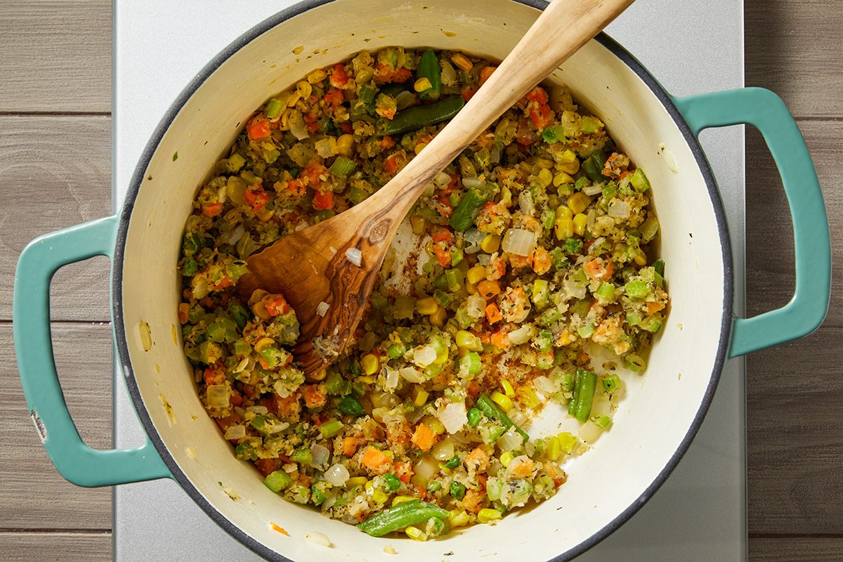 Overhead shot of a large pot with green handles containing a mixture of sautéed vegetables carrots, celery, onions, and green beans with a wooden spoon resting inside, stirring the colorful ingredients