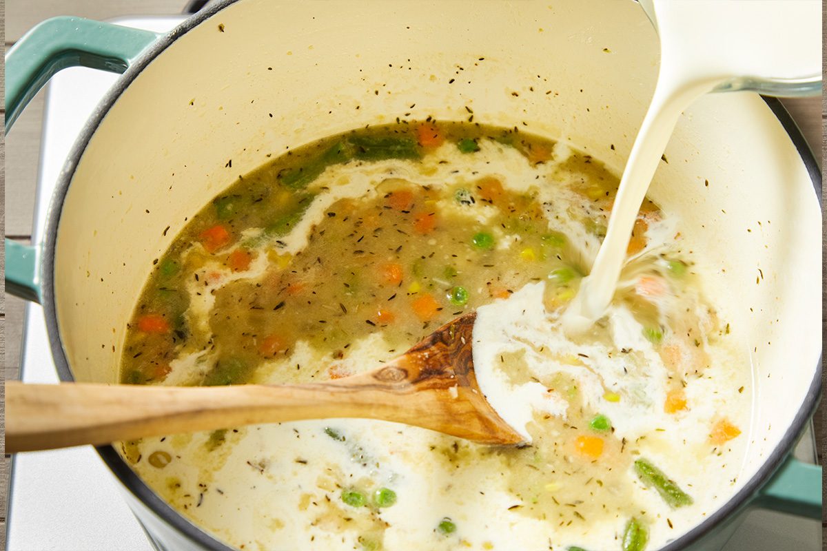 Closeup shot of a wooden spoon stirring a pot of vegetable soup as cream is poured in, creating a creamy swirl over a mix of peas, carrots, and green beans in a light broth
