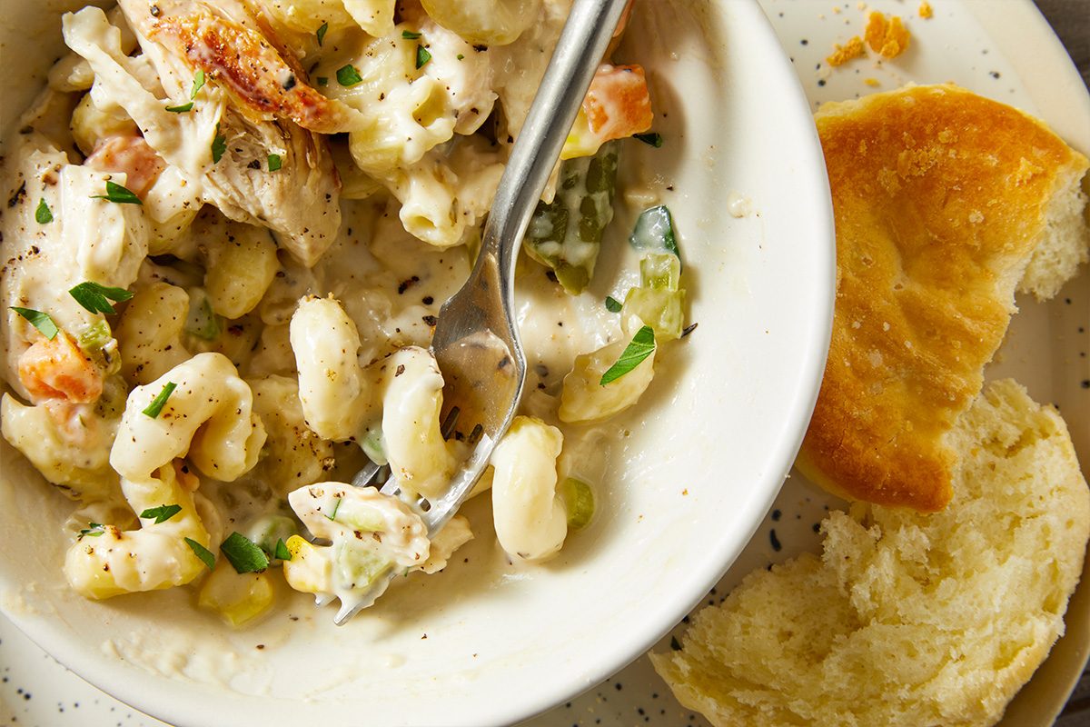 Closeup overhead shot of creamy chicken potpie pasta being scooped with a fork inside a white bowl, showing curls of pasta, chicken, and vegetables; a partially torn biscuit rests on the plate beside the bowl