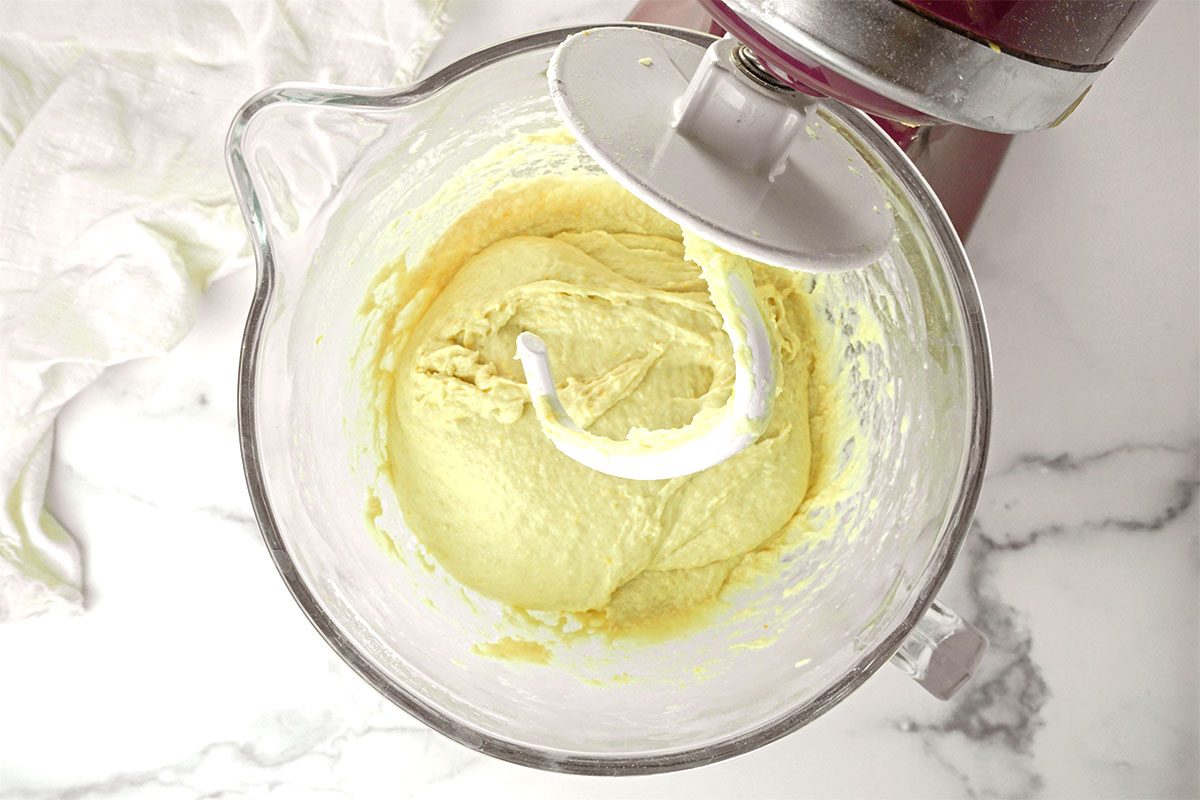 Overhead view of a stand mixer bowl with yellow cake batter being mixed by a white dough hook attachment on a marble countertop. A white cloth is partially visible to the side.