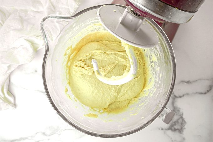 Overhead view of a stand mixer bowl with yellow cake batter being mixed by a white dough hook attachment on a marble countertop. A white cloth is partially visible to the side.