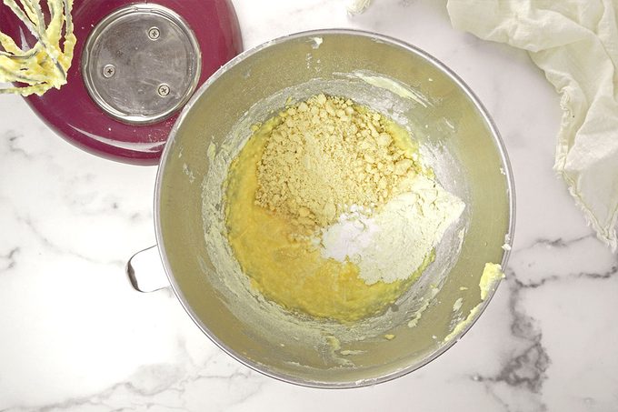 A metal mixing bowl contains partially mixed baking ingredients, including flour, sugar, and possibly baking powder. The bowl is attached to a red stand mixer, with a whisk attachment visible. The background is a white marble surface.