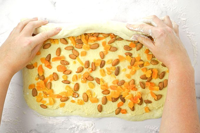 A person rolls up dough that is topped with chopped dried apricots and whole almonds on a floured surface.