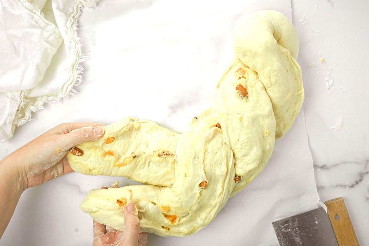 Two hands are braiding a large piece of dough with visible orange bits on a floured white surface. A metal dough scraper and a white cloth are nearby on the countertop.