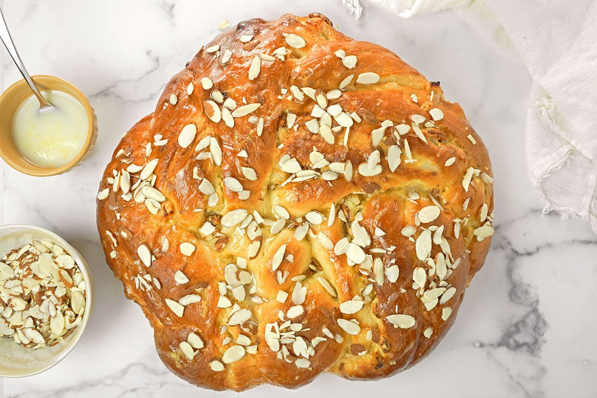 A round, braided loaf of bread topped with sliced almonds sits on a marble surface. Nearby are a small bowl of sliced almonds, a bowl of glaze with a spoon, and a white cloth.