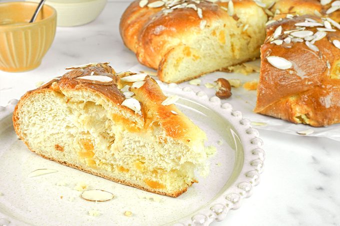 A slice of sweet braided bread with almond slices sits on a plate, with the rest of the loaf and a bowl in the background on a white surface.