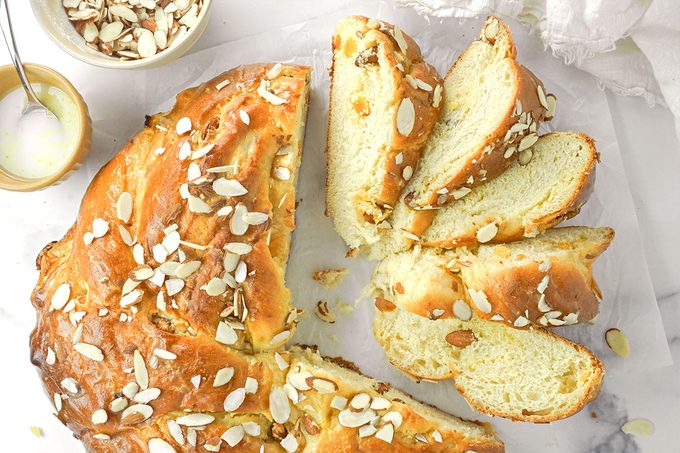A sliced braided bread loaf topped with slivered almonds sits on parchment paper. Nearby are a small bowl of sliced almonds and a bowl with a spoon containing a white glaze.