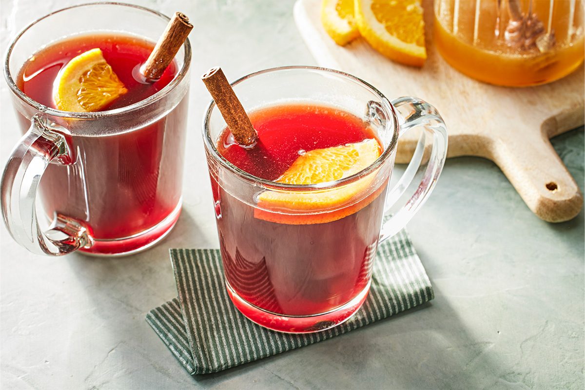 3/4 shot of clear glass mugs filled with deep red Christmas Tea garnished with orange slices and cinnamon sticks, placed on a gray napkin.