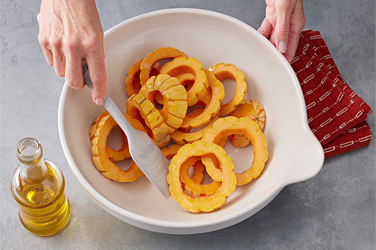 Overhead shot of a person tossing sliced delicata squash with tongs in a white bowl, with a small bottle of olive oil and a red cloth on a gray countertop nearby;