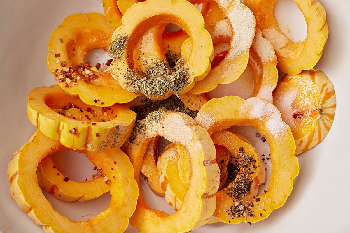 Overhead shot of sliced rings of delicata squash on a white plate, sprinkled with black pepper, salt, dried herbs, and red pepper flakes;