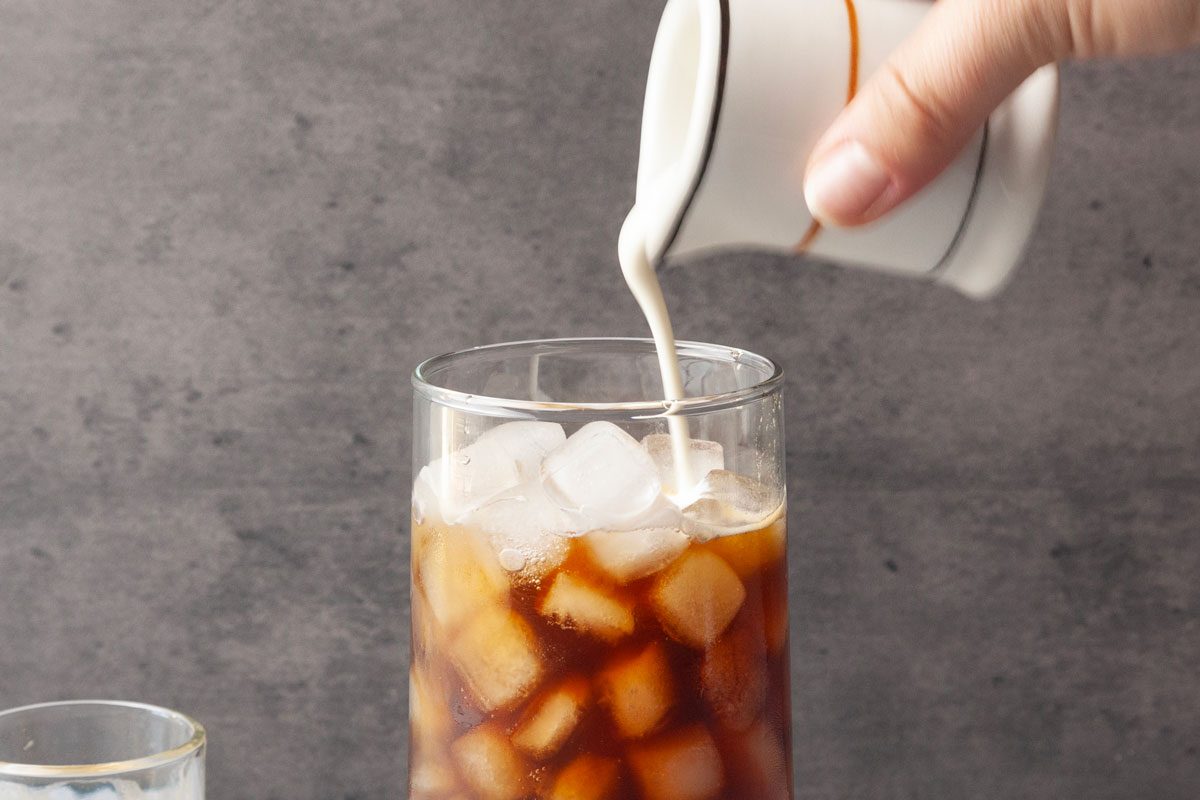 Close shot of a hand pouring cream from a small white pitcher into a tall glass of iced coffee filled with ice cubes, set against a gray background.