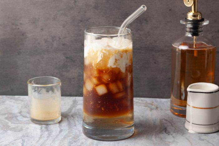 Table view shot of Espresso Cream Soda in a glass with swirling milk and a straw, placed on a marble surface beside a small glass, a pump bottle, and a pitcher.