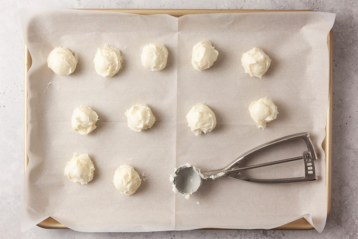 Overhead shot of a parchment-lined baking sheet holding twelve scoops of white cookie dough, with a metal cookie scoop containing a bit of dough resting on the right side of the tray;
