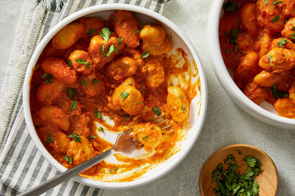 Overhead shot of Indian-Style Butter Gnocchi in a white bowl with tomato sauce and parsley, a fork inside, and extra parsley nearby on a striped cloth;