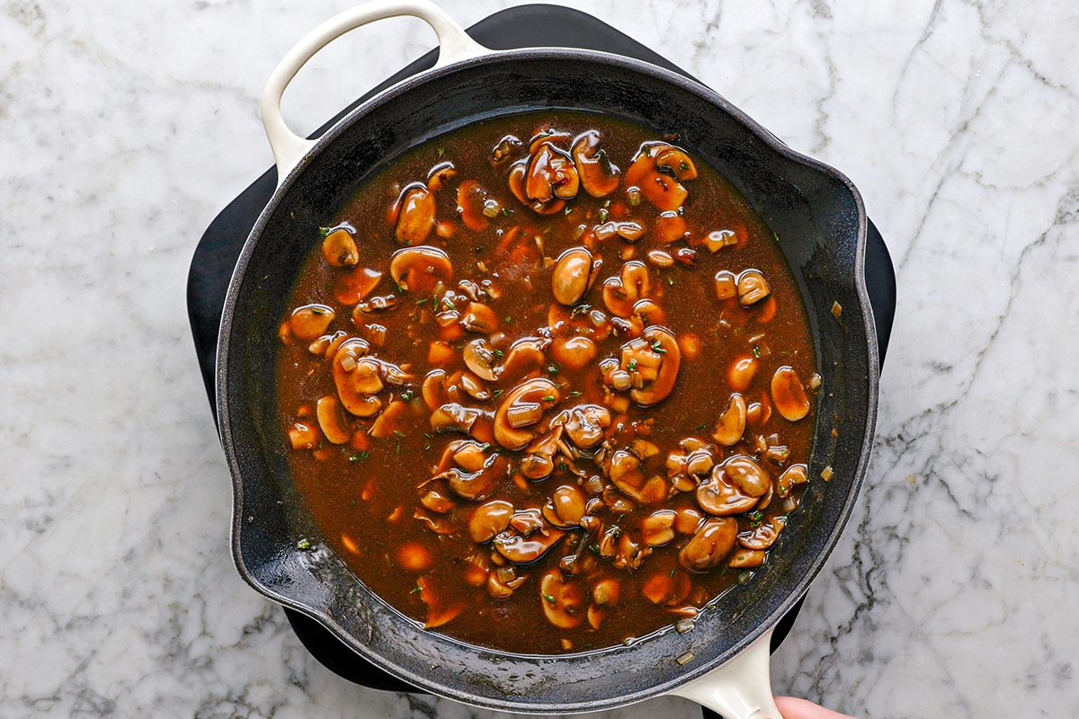 Overhead shot of a skillet filled with brown mushroom sauce on a marble countertop; The sauce contains sliced mushrooms and herbs, with a hand holding the skillet handle in the lower right corner;