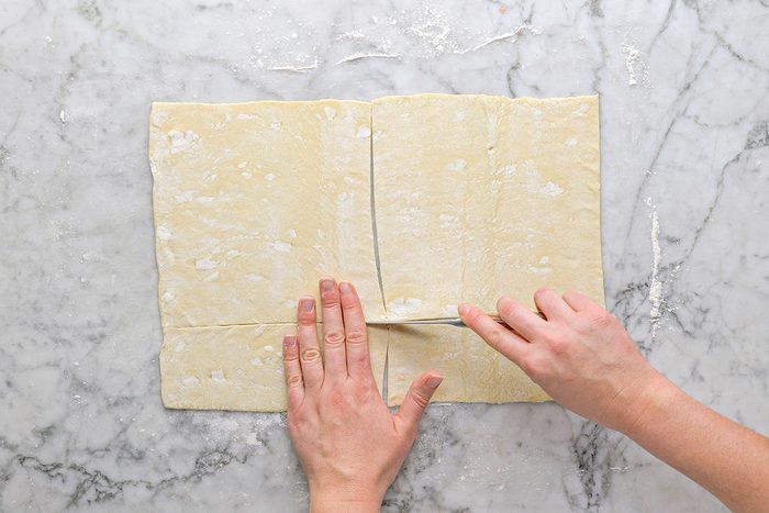Overhead shot of a person’s hands cutting a sheet of puff pastry dough into rectangles on a floured marble surface;