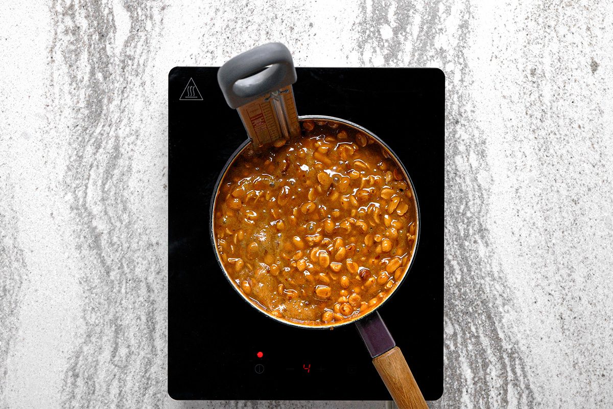 A saucepan filled with beans in a thick, brown sauce is cooking on a black stovetop, with a thermometer clipped to the side of the pot. The surface underneath is a light, marbled countertop.