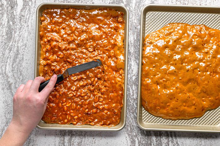 A hand uses an offset spatula to spread peanut brittle mixture evenly on a baking sheet, with another sheet of set peanut brittle next to it on a marble countertop.