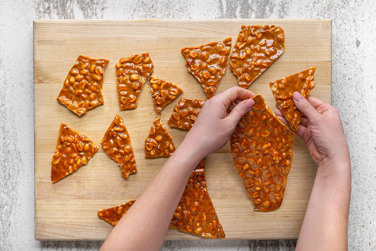 Two hands break a large piece of peanut brittle into smaller shards on a wooden cutting board. Several irregular pieces of the glossy, nut-filled candy are scattered on the board.