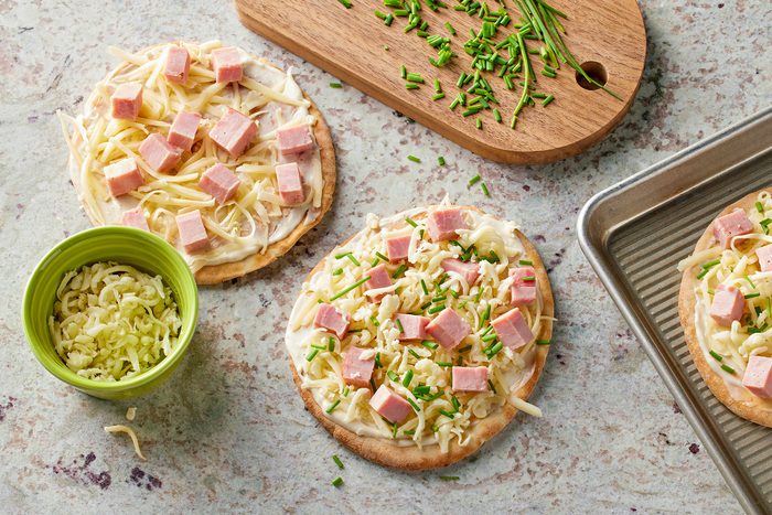 Topping pita bread with Swiss cheese, ham and mozzarella cheese, then sprinkling them with chives. cheese and chives on side.