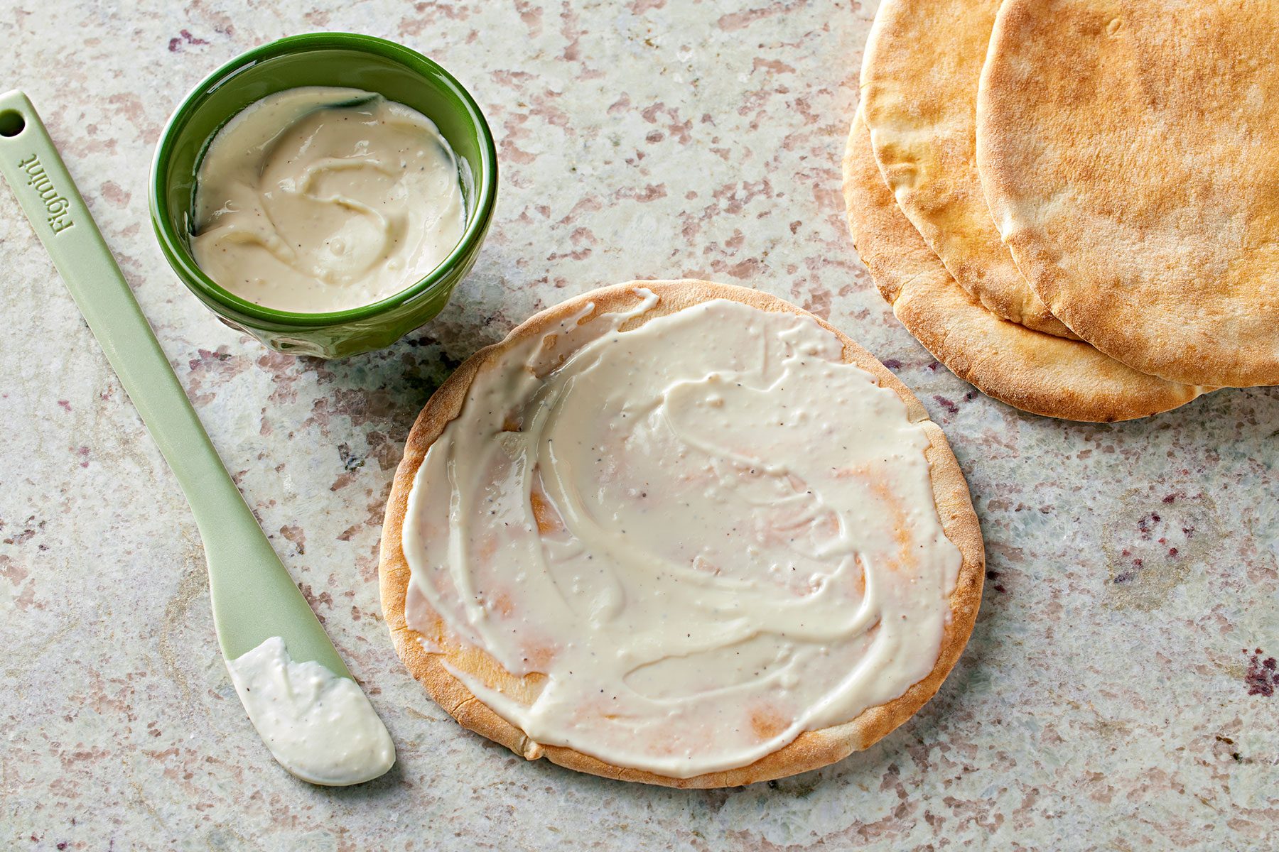 Spreading Alfredo sauce evenly over the pita breads, leaving a small border around the edge for a crust.