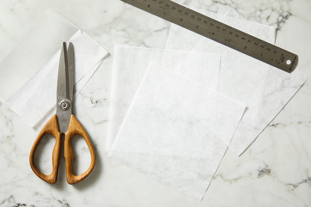 Overhead shot of a pair of scissors with wooden handles, a metal ruler, and several sheets of white wax or parchment paper arranged on a white marble surface.