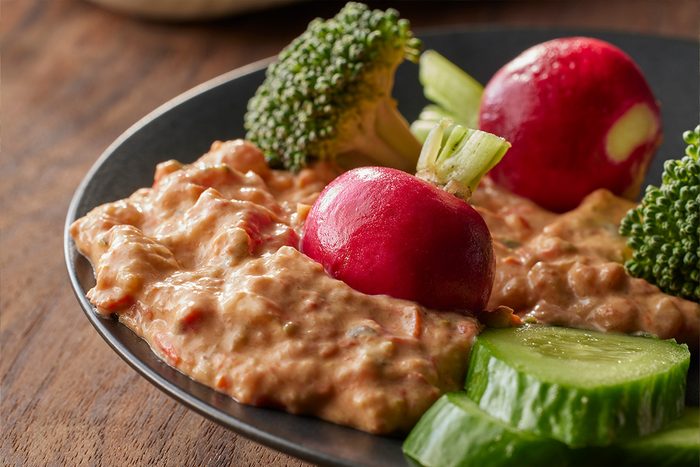 Close-up of Roasted Vegetable Dip served on a dark dish, accompanied by two red radishes, broccoli florets, and cucumber slices;