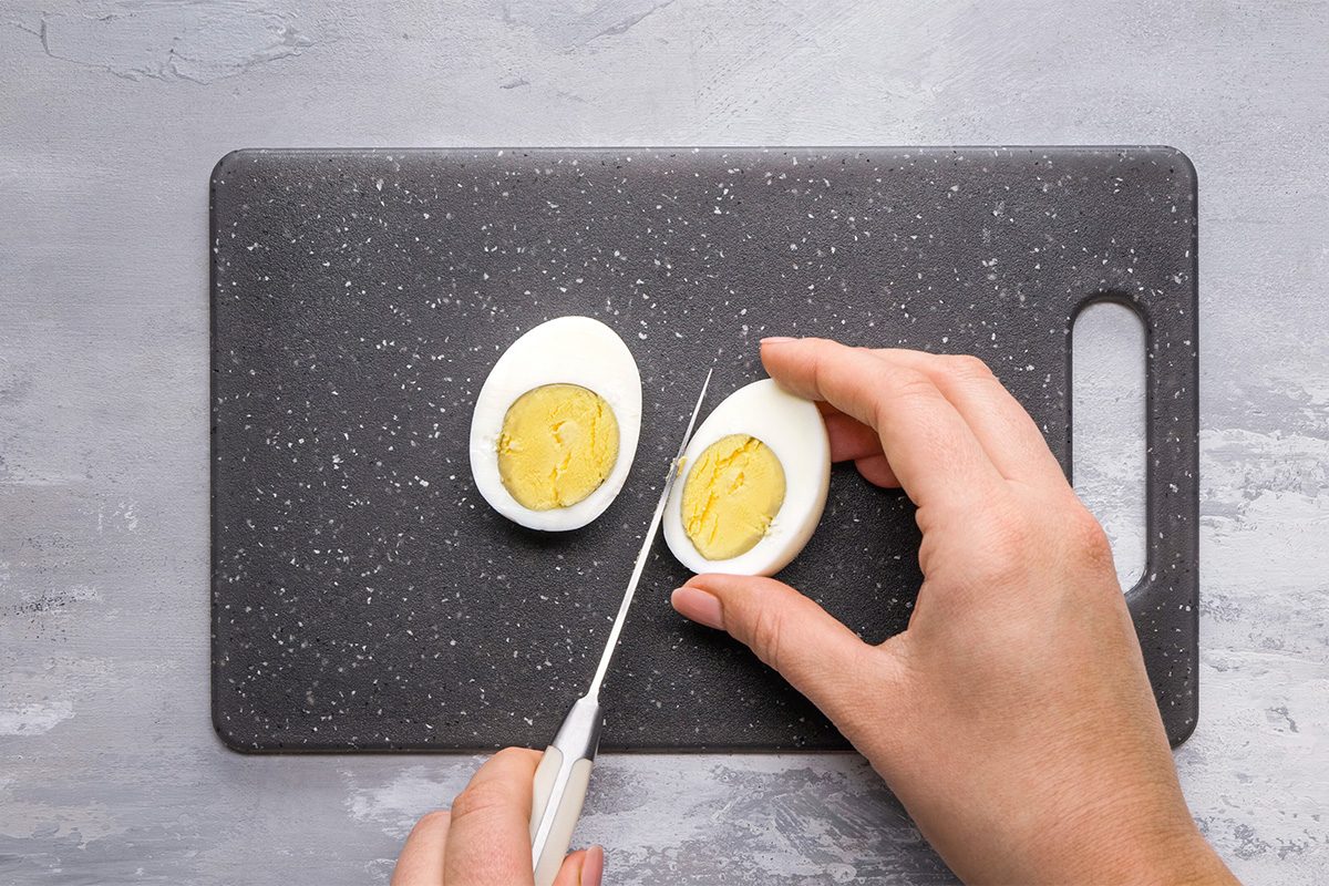 A person slices a hard-boiled egg in half on a black cutting board with a white-handled knife. The egg yolk and egg white are clearly visible.