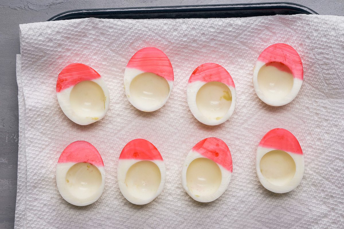 Eight hard-boiled egg halves with the tops painted red are arranged on a paper towel-lined tray. The eggs are evenly spaced and the red color covers the top portion of each egg half.