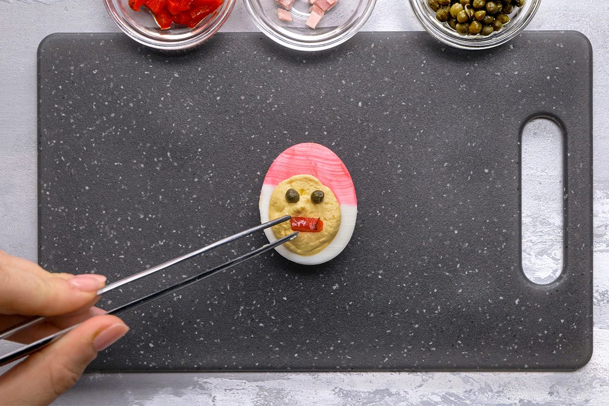 A hand uses tweezers to add a red pepper strip to a boiled egg decorated with capers and other toppings to resemble a face, on a black cutting board with small bowls of ingredients nearby.