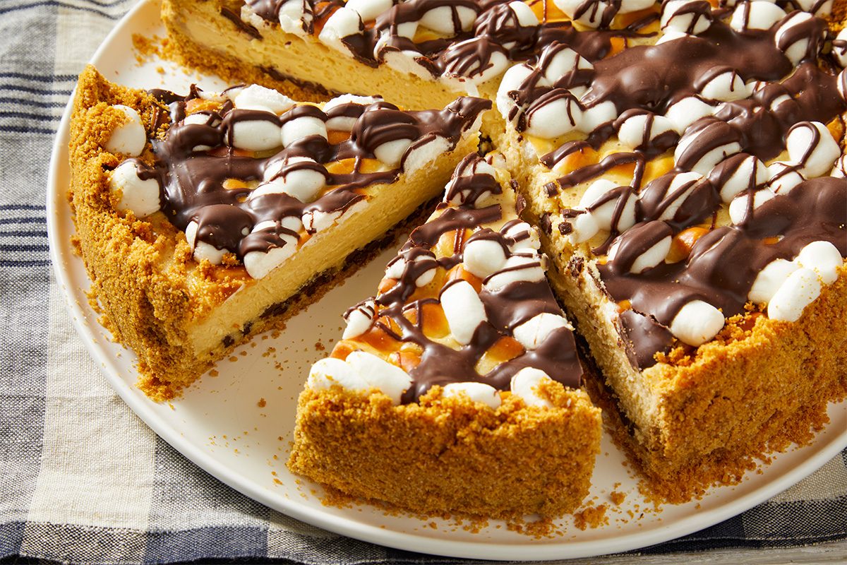 A sliced cheesecake with a graham cracker crust, topped with mini marshmallows, chocolate drizzle, and caramel sauce, sits on a white plate over a plaid tablecloth.