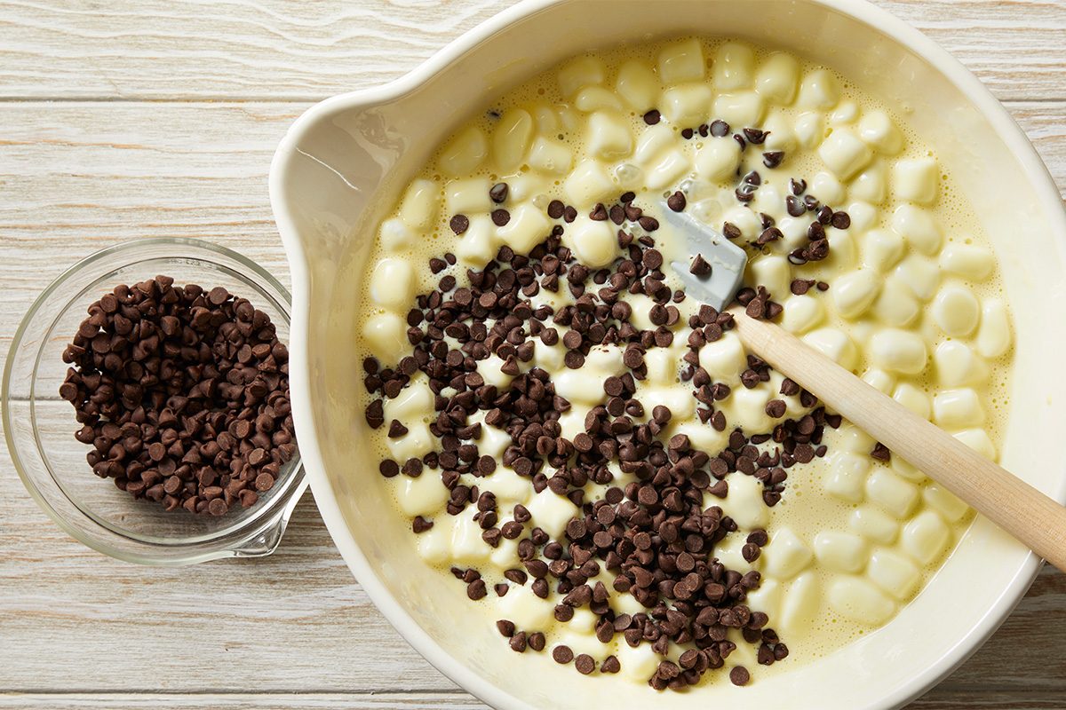 A mixing bowl filled with melted marshmallows and chocolate chips being stirred with a spatula, next to a small glass bowl of extra chocolate chips on a light wooden surface.
