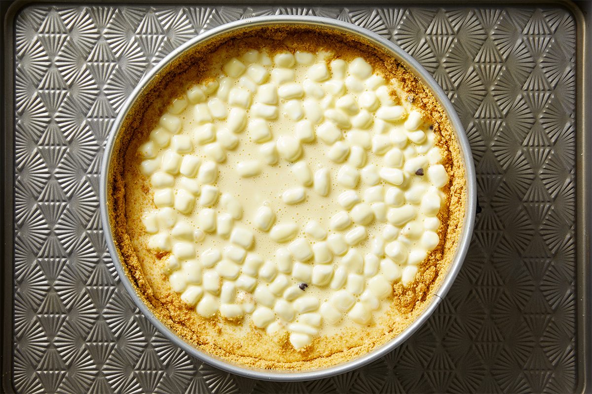 A round pie with a golden graham cracker crust, topped with a layer of mini marshmallows, sits on a textured, patterned metal baking sheet.