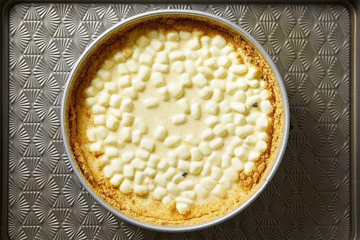 A round pie with a golden graham cracker crust, topped with a layer of mini marshmallows, sits on a textured, patterned metal baking sheet.