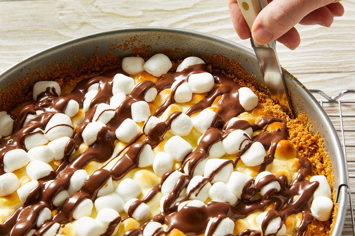 A hand slices into a pie topped with mini marshmallows and drizzled chocolate, set on a graham cracker crust in a round pan on a wooden surface.