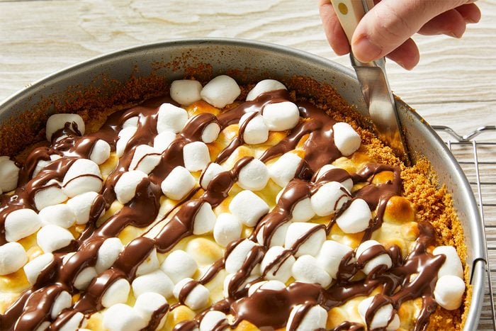 A hand slices into a pie topped with mini marshmallows and drizzled chocolate, set on a graham cracker crust in a round pan on a wooden surface.