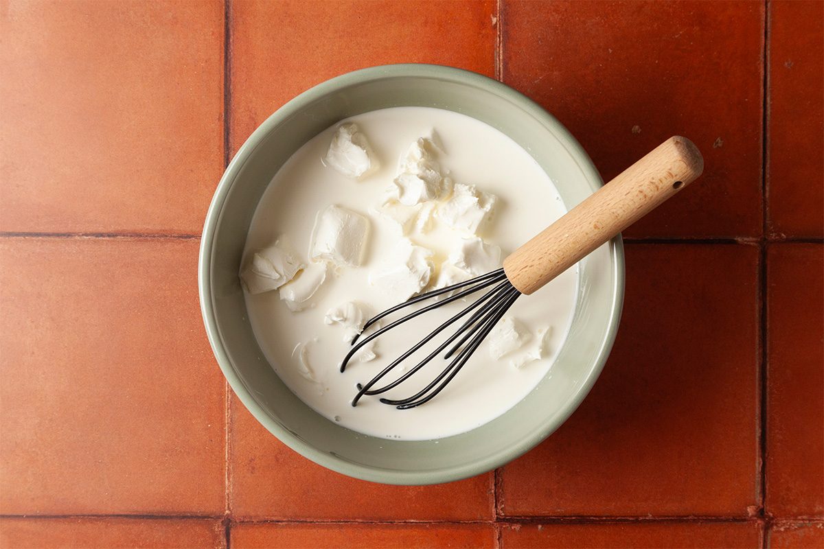 Overhead shot of a bowl containing milk and chunks of white cheese or cream, with a whisk featuring a wooden handle resting inside, placed on a red tiled surface;