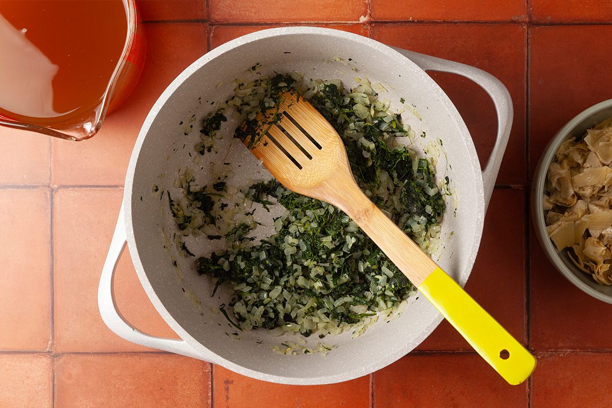 Overhead shot of a white pot with chopped onions and herbs being sautéed, stirred with a wooden spoon with a yellow handle, on a red tiled surface; Nearby are a bowl of ingredients and a measuring cup with liquid
