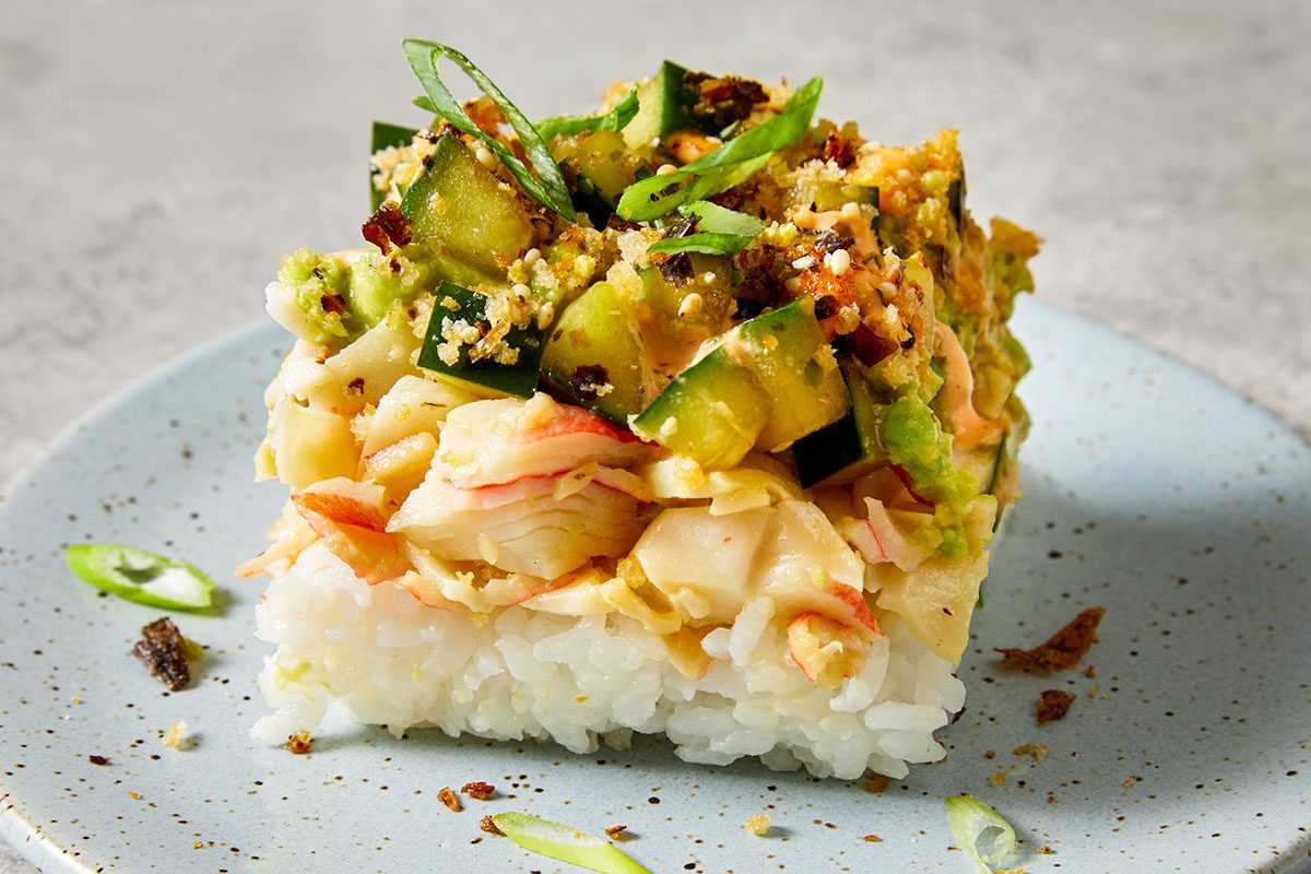 Closeup shot of a single square serving of Sushi Bake; layered with fluffy white rice, shredded imitation crab, creamy toppings, diced cucumber, toasted breadcrumbs, and sliced green onions; presented on a light blue speckled plate;