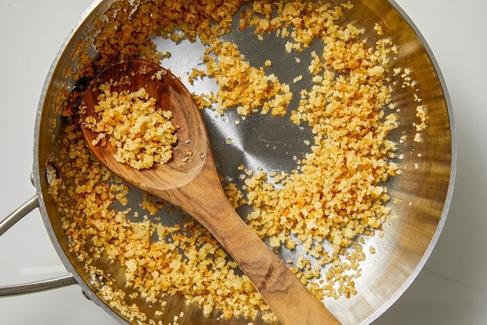 Overhead shot of a stainless steel pan filled with golden-brown breadcrumbs; a wooden spoon rests inside; toasted crumbs are scattered across the bottom of the pan;