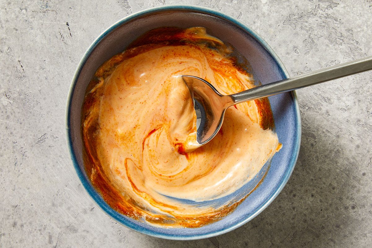 Overhead shot of a blue bowl filled with a creamy orange-and-white sauce; a silver spoon is stirring the mixture; set on a textured gray surface;