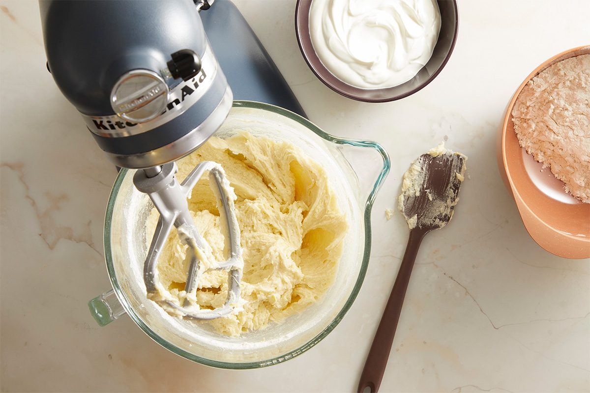 A stand mixer with creamy batter in a glass bowl sits on a countertop next to a spatula with batter, a bowl of white cream, and a bowl of dry ingredients.