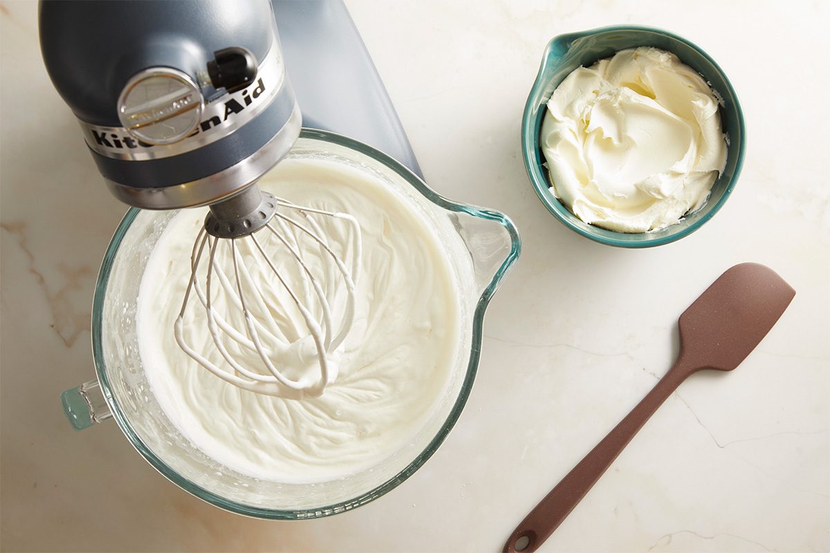 An electric stand mixer with a whisk attachment is whipping a creamy mixture in a glass bowl. Nearby, a bowl of cream cheese and a brown spatula rest on a light countertop.